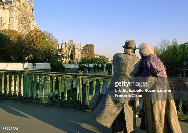 france, paris, elderly couple walking arm in arm on bridge next to notre dame cathedral - quayside stock pictures, royalty-free photos & images