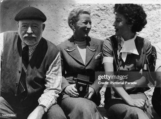 American writer Ernest Hemingway smiles at the camera while his fouth wife, journalist Mary Welsh Hemingway and actress Ava Gardner sit nearby and...