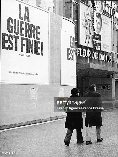 Arm in arm, a couple walk past posters that read 'La Guerre est Finie! Si Vous le Voulez. John et Yoko Lennon vous souhaitent un Joyeux Noel,' on the...