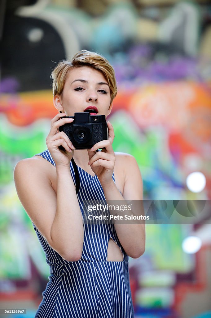 Portrait of cool teenage girl holding up camera in front of graffiti wall