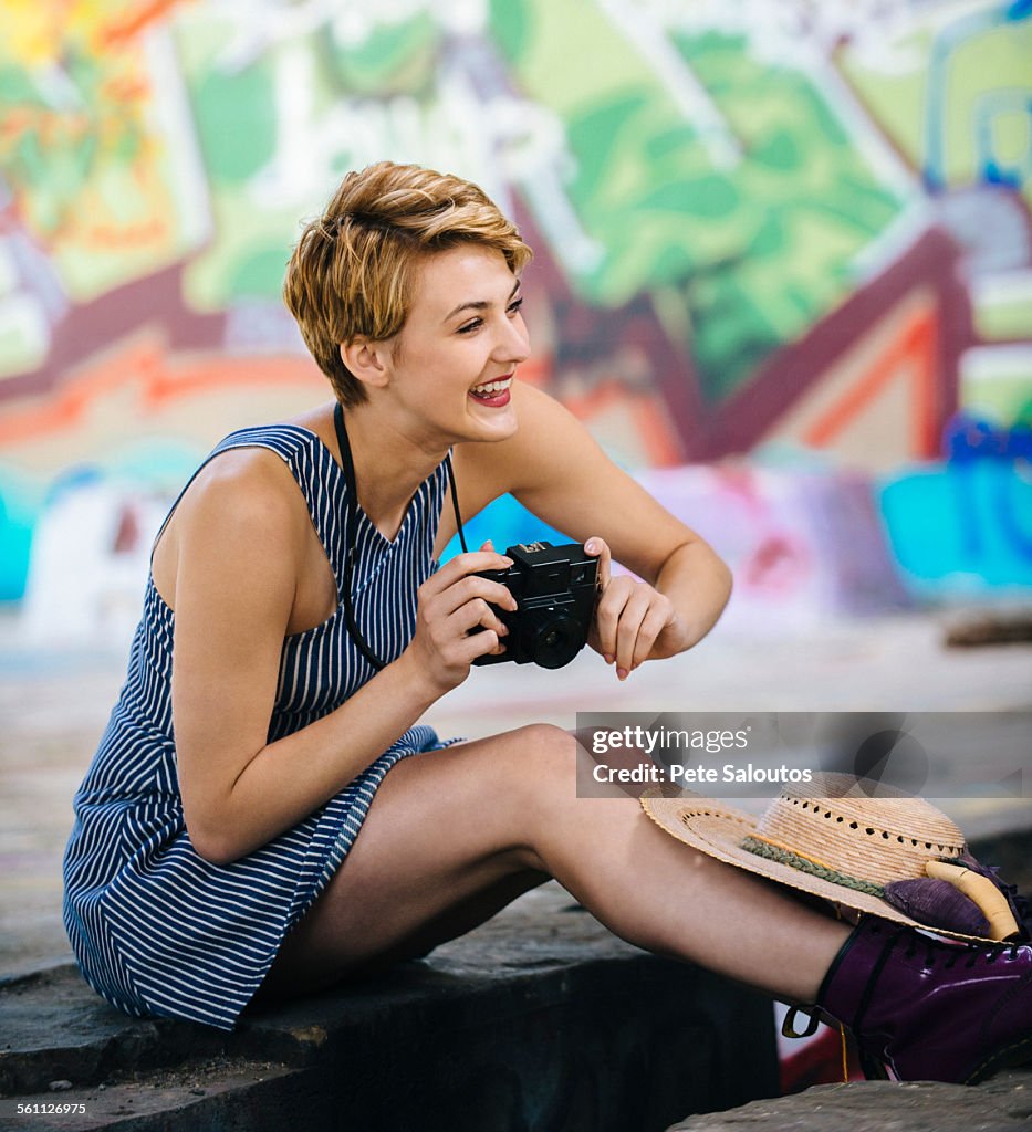 Stylish teenage girl sitting on sidewalk with camera in front of graffiti wall
