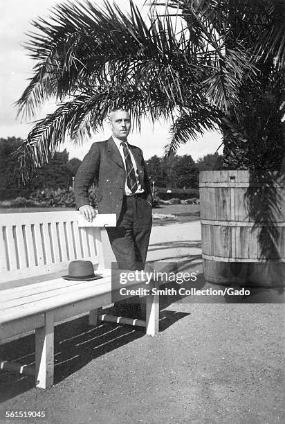 Man in a suit with a hat leaning on an outdoor bench, under a palm tree, Germany, 1950.