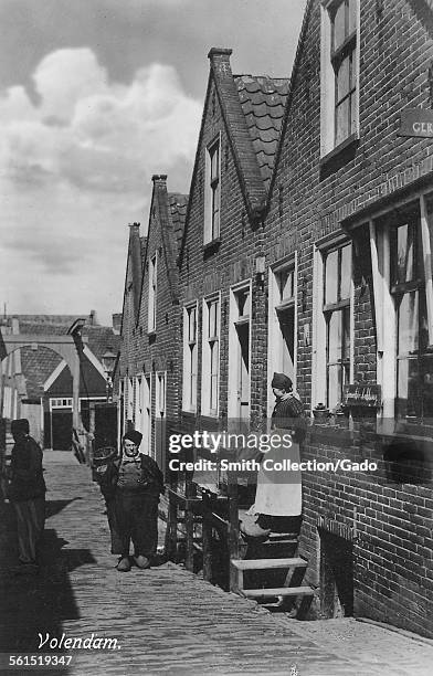 Street life, a woman standing on her doorstep and a man walking along narrow cobbled street and a canal bridge in the distance, Volendam,...