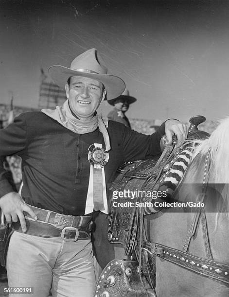 Actor John Wayne, wearing a cowboy hat, with hands on hips, standing with a horse and a medal, 1954.