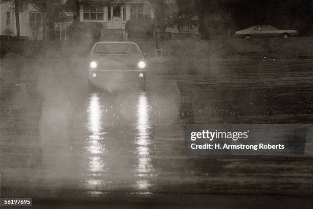 1970s: Automobile Car With Headlights On, Coming Down Rainy Street At Night In Bad Weather, Showing Rain, Cars And Lights.