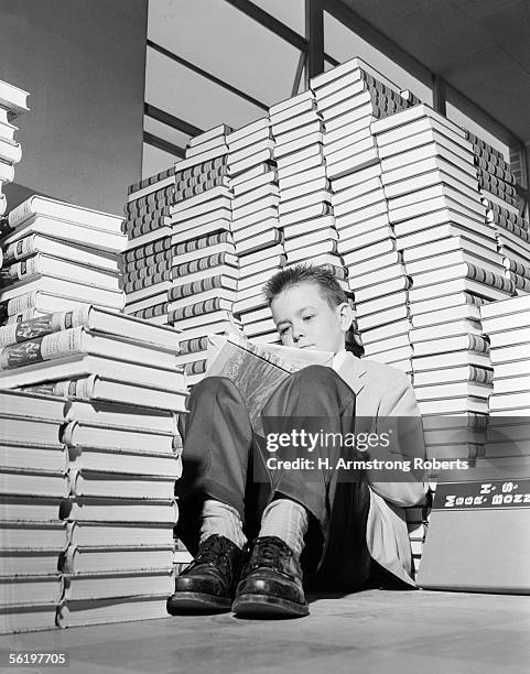 1950s: Books Are Piled High And Surround A Boy, Sitting On The Floor With Knees Up And Reading A Book. He Is Dressed In Sport Jacket And Slacks, With...