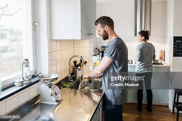 man washing sauce pan while woman standing in background in kitchen - man housework stock pictures, royalty-free photos & images