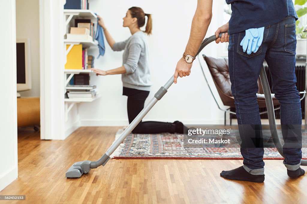 Low section of man vacuuming floor while woman cleaning shelves in background at home