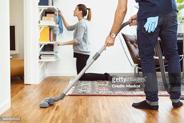 low section of man vacuuming floor while woman cleaning shelves in background at home - low section woman stock pictures, royalty-free photos & images