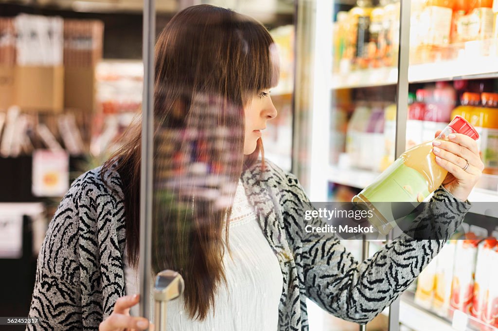 Young woman reading label on juice bottle at refrigerated section in supermarket