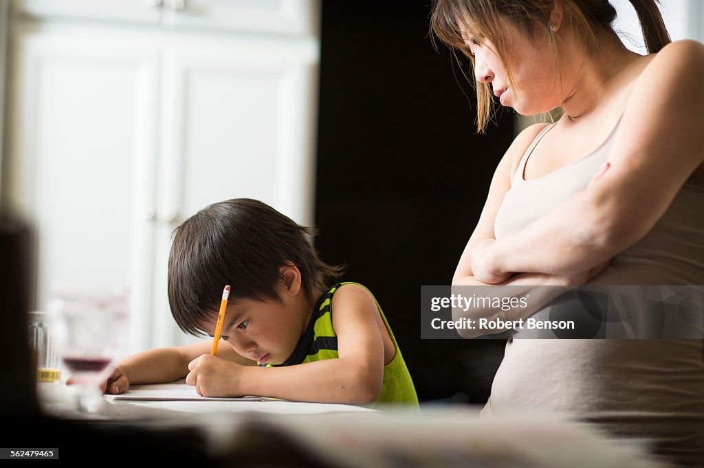 A Japanese boy studies Japanese homework in a kitchen as his mom looks on.