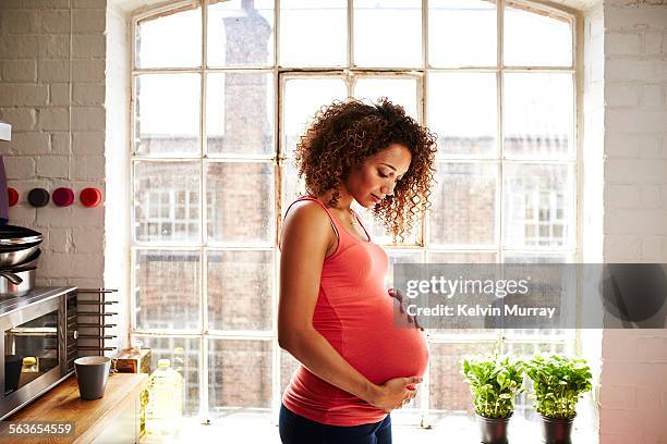 a pregnant woman holds her bump in kitchen window - gravid imagens e fotografias de stock