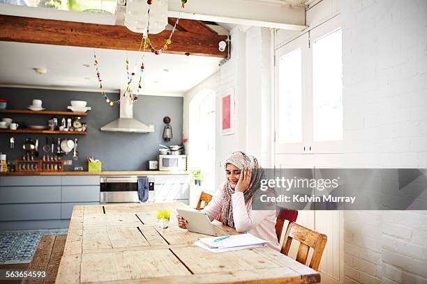 woman uses tablet while sitting at a table - headscarf home stockfoto's en -beelden