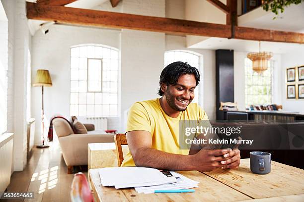 a man smiles whilst using his phone at home - paperwork stock pictures, royalty-free photos & images