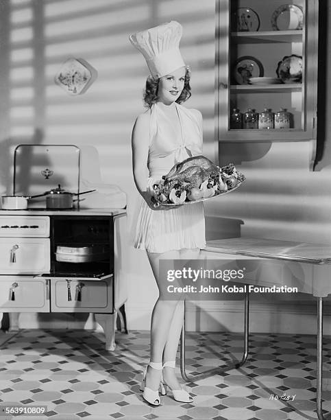 Portrait of actress Arlene Dahl holding a roast chicken and wearing a chef hat, for Warner Bros Studios, 1946.