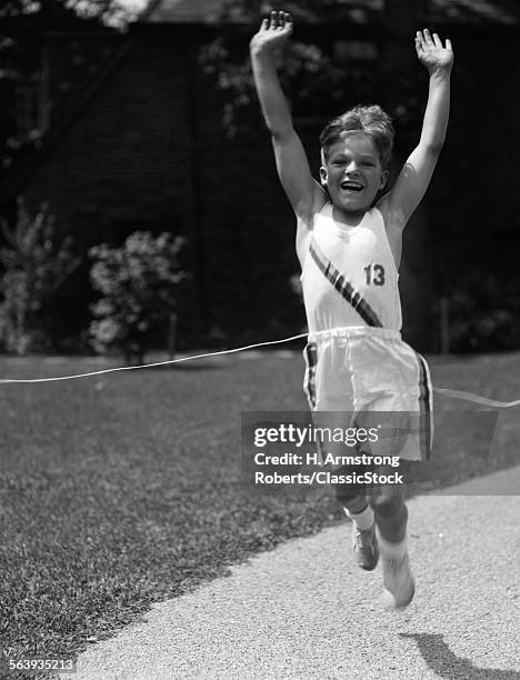 1930s BOY IN TRACK UNIFORM WITH ARMS IN AIR BREAKING TAPE AT FINISH LINE LOOKING AT CAMERA