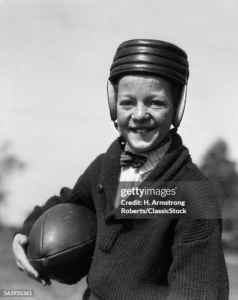 1920s FRECKLE-FACED BOY IN SWEATER & BOW TIE WEARING LEATHER FOOTBALL HELMET HOLDING FOOTBALL UNDER ARM LOOKING AT CAMERA