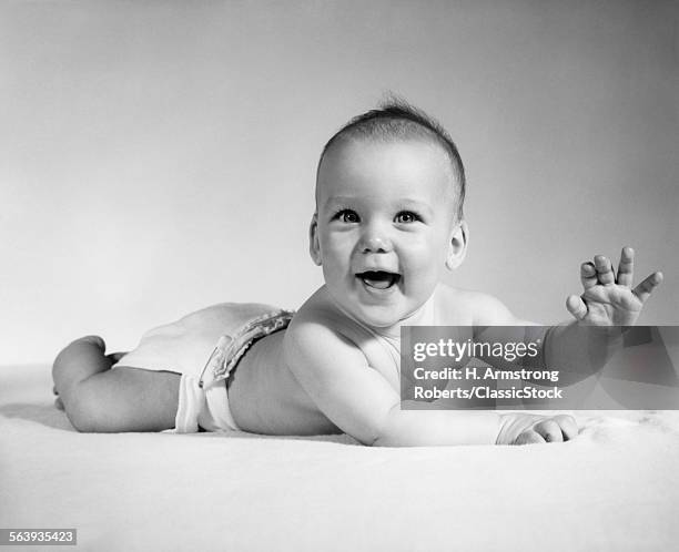 1960s SMILING BABY WEARING CLOTH DIAPER LYING ON STOMACH LOOKING AT CAMERA ARM RAISED