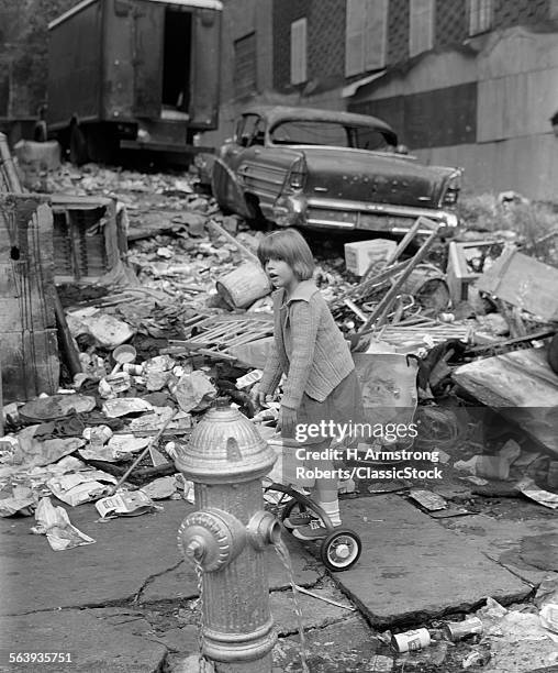 1960s GIRL STANDING ON BACK OF TRICYCLE BEHIND OPEN FIRE HYDRANT SURROUNDED BY WRECKAGE WITH OLD BEAT-UP CAR IN BACKGROUND