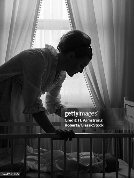 1960s SILHOUETTE OF MOTHER IN NIGHTGOWN CHECKING IN ON BABY LYING IN CRIB WITH SUNLIGHT PEEKING IN THROUGH WINDOW IN BACKGROUND