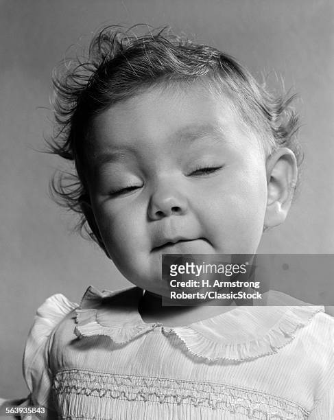 1950s PORTRAIT BABY IN FRILLY DRESS WITH MESSY HAIR CLOSED EYES FUNNY FACIAL EXPRESSION