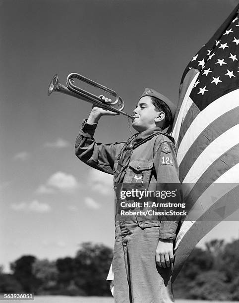 1950s PROFILE OF BOY SCOUT IN UNIFORM STANDING IN FRONT OF 48 STAR AMERICAN FLAG BLOWING BUGLE