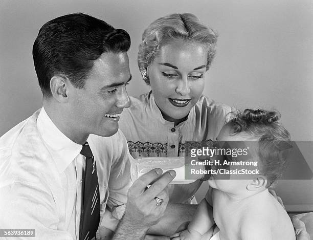 1950s SMILING FATHER FEEDING BABY MILK FROM BOTTLE WITH MOTHER AT SIDE