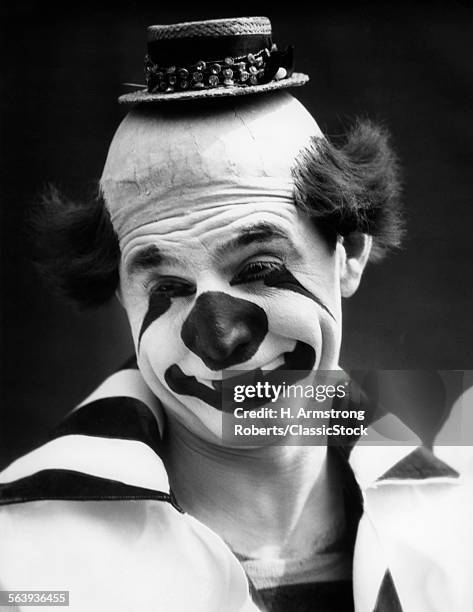 1930s CLOWN IN CLASSIC WHITEFACE WITH A HAPPY SAD FACE WITH A BALD HEAD WEARING A TINY STRAW HAT LOOKING AT CAMERA