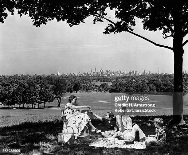 1930s 1940s FAMILY PICNICKING UNDER A TREE IN FAIRMONT PARK WITH SKYLINE OF PHILADELPHIA PA ON HORIZON