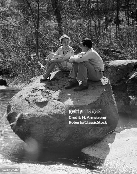 1930s COUPLE MAN WOMAN SITTING ON LARGE ROCK IN STREAM HUGGING KNEES SMILING HAPPY ROMANCE