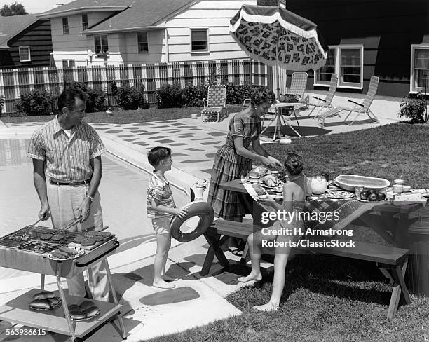 1960s FAMILY OF 4 IN BACKYARD AT POOL SIDE FATHER BARBECUING & MOTHER & CHILDREN MAKING PREPARATIONS AT PICNIC TABLE