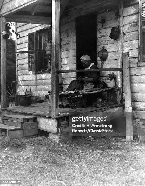 1920s 1930s ELDERLY AFRICAN AMERICAN WOMAN SITTING ON PORCH OF SMALL WOODEN HOUSE