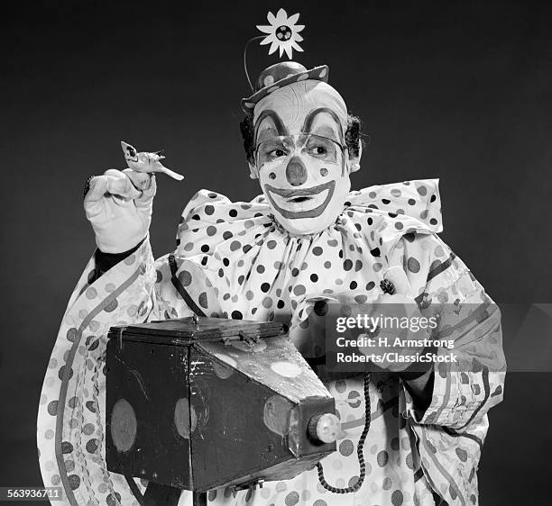 1960s CLOWN IN POLKA-DOTTED OUTFIT TAKING PICTURE WITH OLD-TIME CAMERA HOLDING BIRDIE IN OPPOSITE HAND