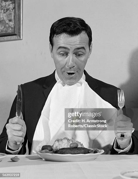 1960s MAN SITTING AT TABLE READY TO EAT DINNER WITH NAPKIN AT NECK AND KNIFE & FORK IN AIR