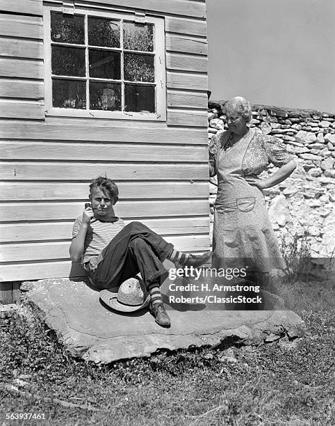 1940s FARM BOY LEANING AGAINST BUILDING SMOKING PIPE WITH GRANDMOTHER LOOKING AROUND CORNER AT HIM WITH HAND ON HIP