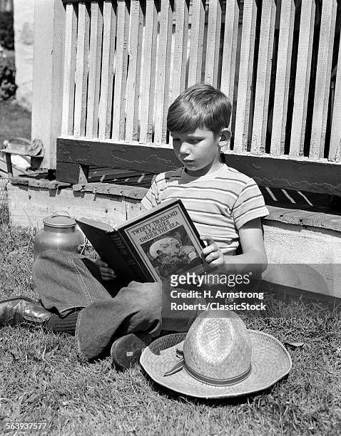 1940s BOY IN JEANS & STRIPED T-SHIRT LEANING AGAINST PORCH READING THE BOOK TWENTY THOUSAND LEAGUES UNDER THE SEA