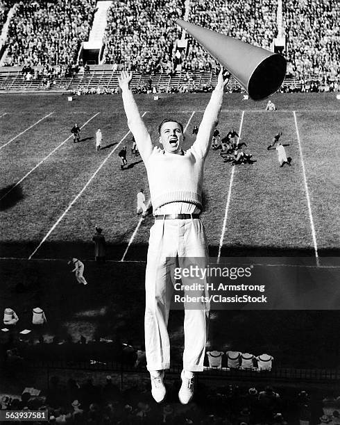 1940s MALE CHEERLEADER WITH MEGAPHONE JUMPING IN AIR AT FOOTBALL GAME