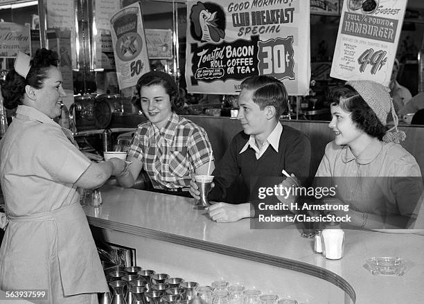 1950s SMILING WOMAN WAITRESS SERVING TWO TEENAGE GIRLS AND ONE BOY AT DINER COUNTER