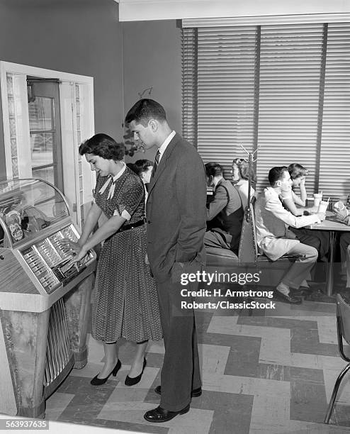 1950s TEEN COUPLE PLAYING JUKE BOX IN MALT SHOP WITH OTHER TEENS IN BOOTHS