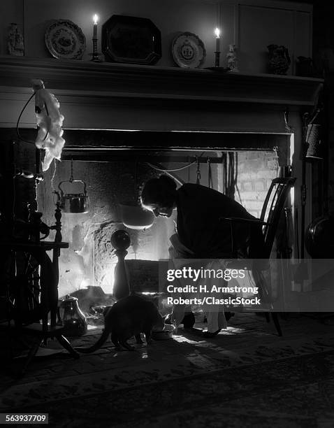 1920s WOMAN SITTING BY FIREPLACE IN CHAIR LEANING OVER PLACING BOWL TO FEED CAT ON FLOOR ONLY LIGHT FROM FIRE & CANDLES