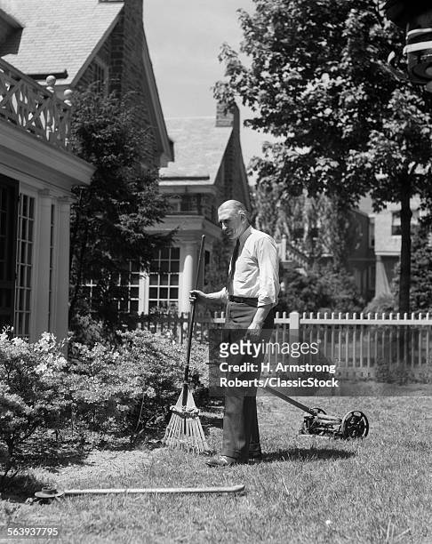 1930s SENIOR MAN STANDING BY GARDEN SHRUBS WITH RAKE PUSH MOWER LAWNMOWER BEHIND HIM