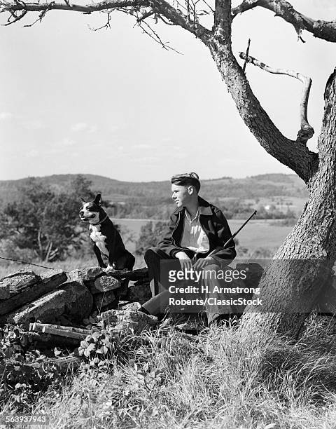 1930s BOY SITTING ON STONE FENCE WITH BOSTON TERRIER DOG RESTING RIFLE BETWEEN LEGS