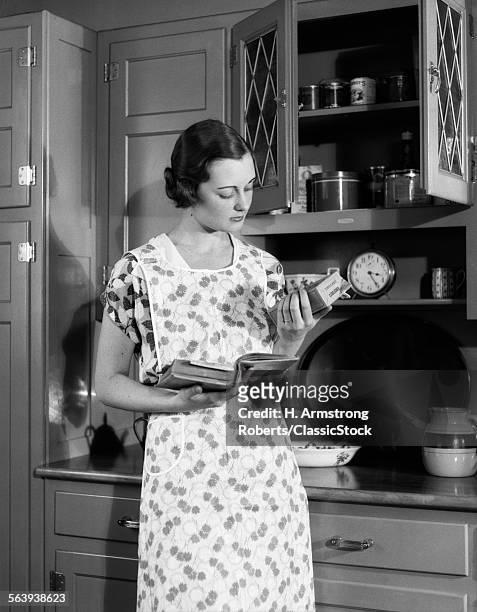 1920s 1930s WOMAN HOUSEWIFE WEARING APRON IN KITCHEN HOLDING COOKBOOK READING PACKAGE OF CORN STARCH
