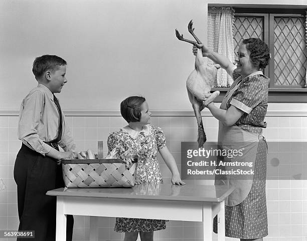 1940s HOUSEWIFE IN KITCHEN SHOWING RAW FRESH PLUCKED TURKEY TO SON AND DAUGHTER