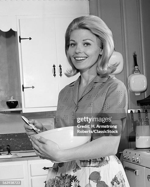 1960s 1970s SMILING BLOND WOMAN HOUSEWIFE IN KITCHEN HOLDING MIXING BOWL AND SPOON LOOKING AT CAMERA