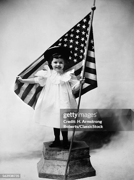 1890s TURN OF CENTURY SMILING LITTLE GIRL STANDING ON PLATFORM WEARING A WHITE DRESS AND A DARK HAT HOLDING A AMERICAN FLAG