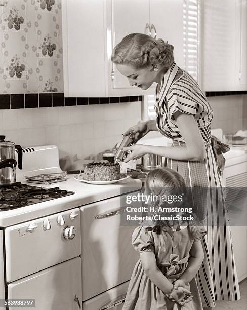 1950s HOUSEWIFE IN KITCHEN DECORATING CAKE ON STOVE WITH PASTRY GUN WHILE LITTLE DAUGHTER WATCHES