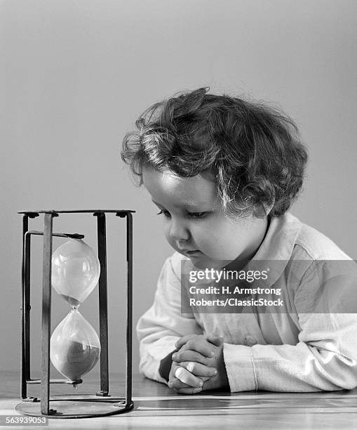 1940s YOUNG GIRL TODDLER LEANING ELBOWS ON TABLE STARING AT HOUR GLASS CURIOSITY SAND TIME ETERNITY