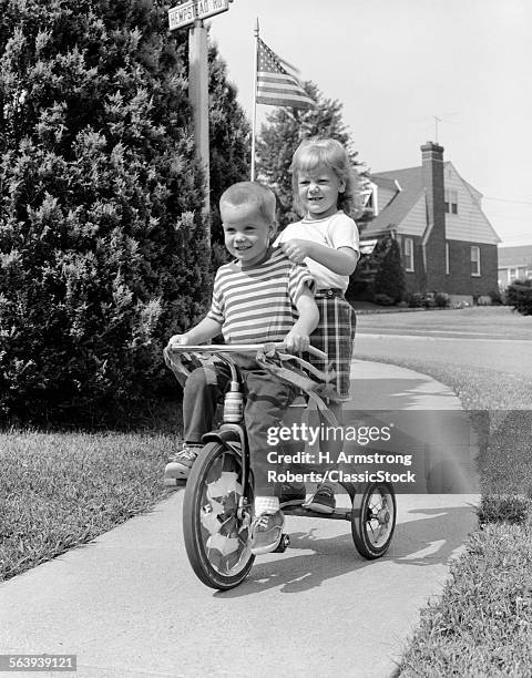 1960s TWO CHILDREN RIDING TRICYCLE ON SUBURBAN SIDEWALK