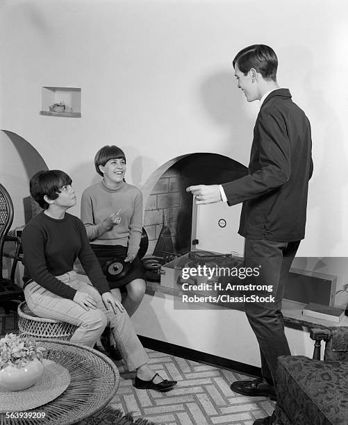 1960s 2 TEEN GIRLS SEATED ON WICKER STOOL AT FIREPLACE CHANGING 45 RPM RECORDS WITH TEEN BOY DANCING